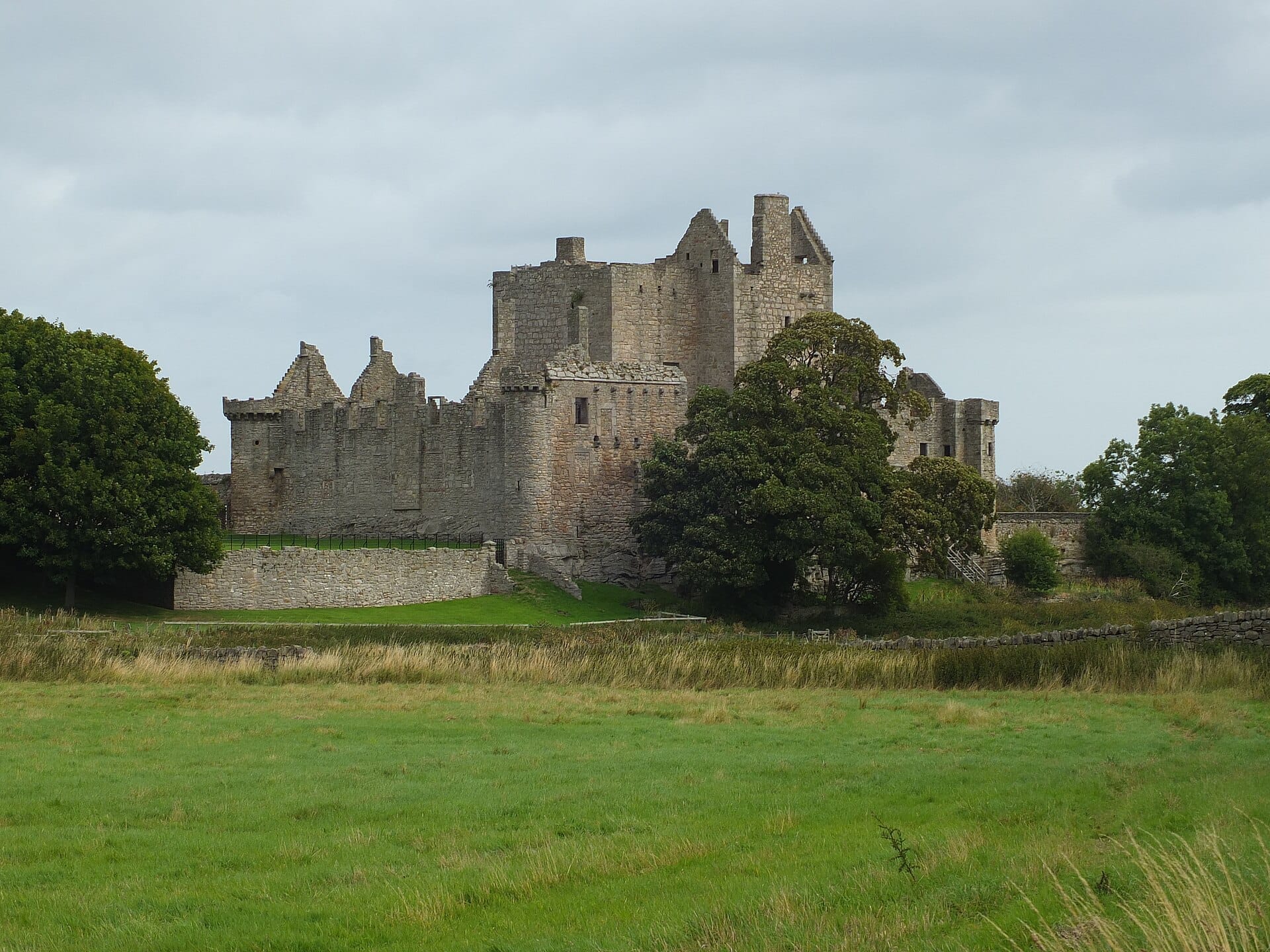 Craigmillar Castle, eine mittelalterliche Steinfestung, steht auf einem grasbewachsenen Hügel nahe Edinburgh, umgeben von Bäumen unter einem bewölkten Himmel.