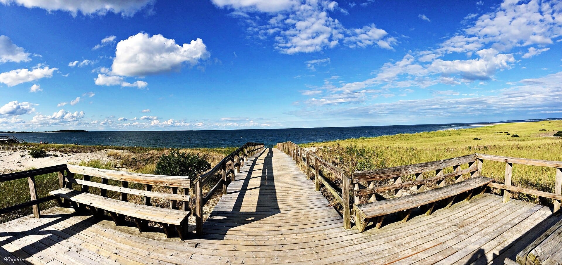 Ampia passerella in legno e panchine che si affacciano sulle dune di sabbia e sull'acqua azzurra a Crane Beach, con paesaggio erboso sotto un cielo luminoso.