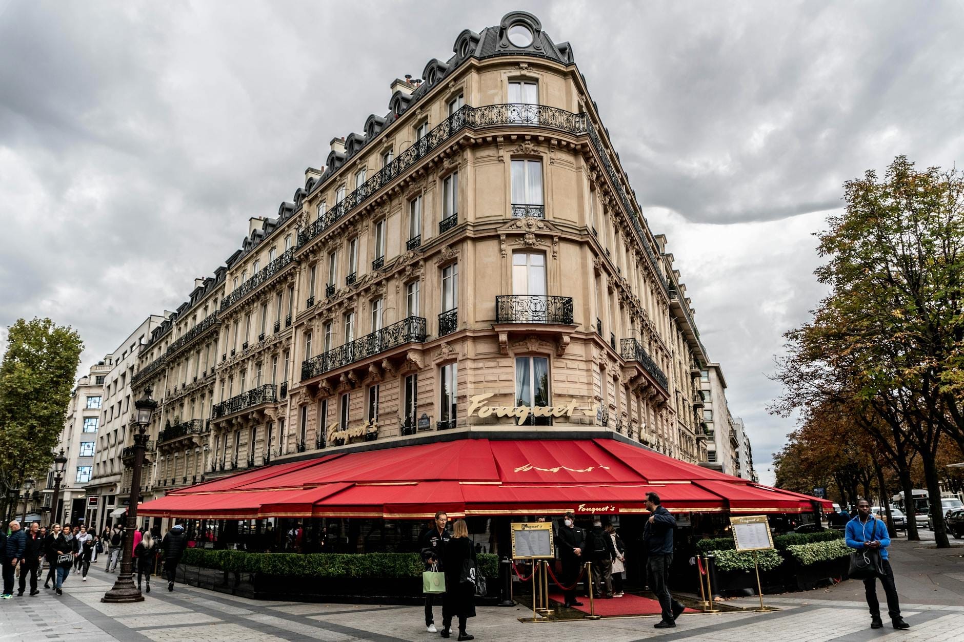 Elegante edificio parigino su un angolo animato con una tenda rossa, persone che si radunano all'esterno e alberi che fiancheggiano la strada sotto un cielo nuvoloso e drammatico.