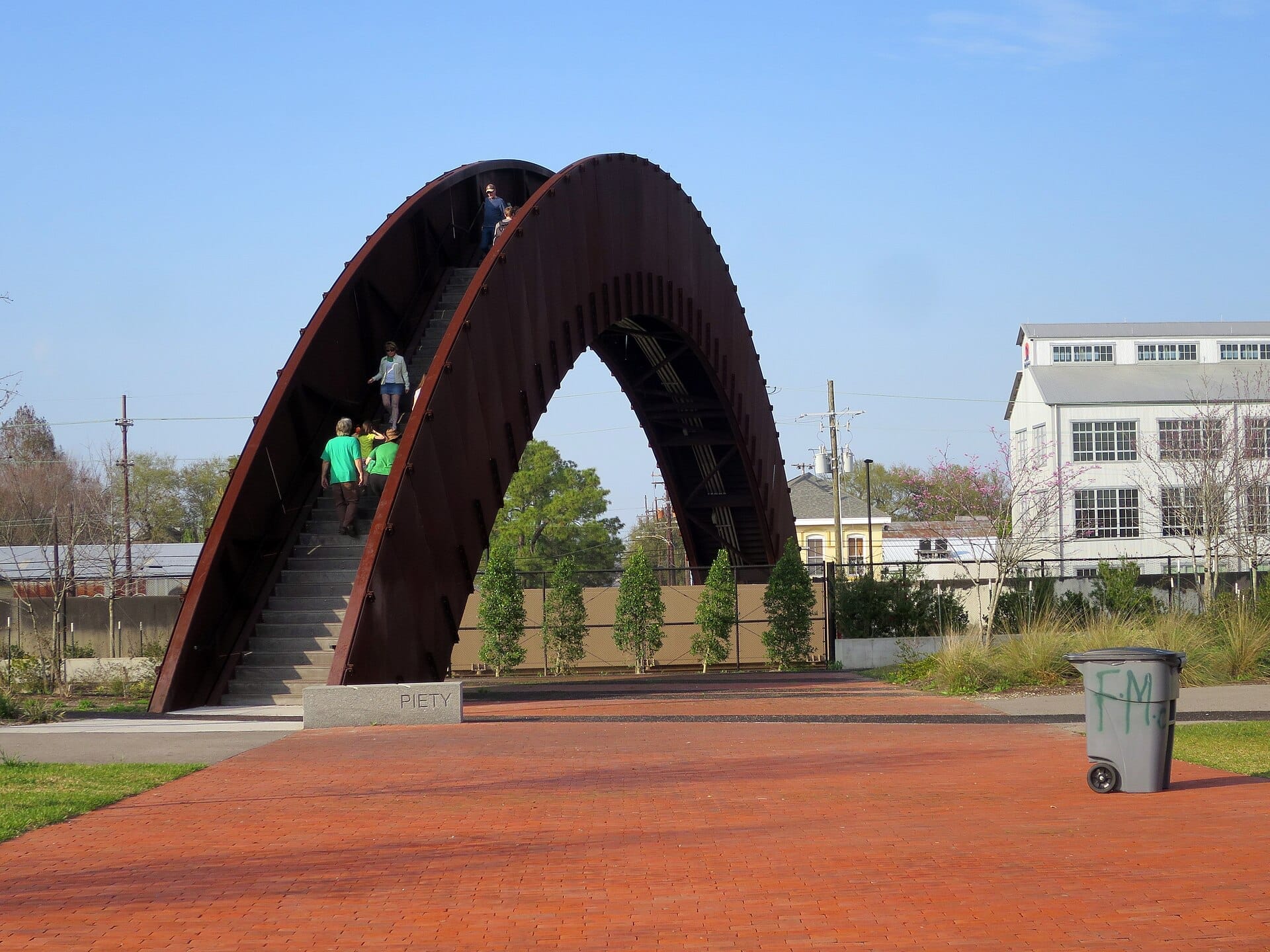Passerella pedonale al Crescent Park con persone che salgono le scale ad arco, circondata da vegetazione e dagli edifici del quartiere Bywater sotto un cielo azzurro sereno.