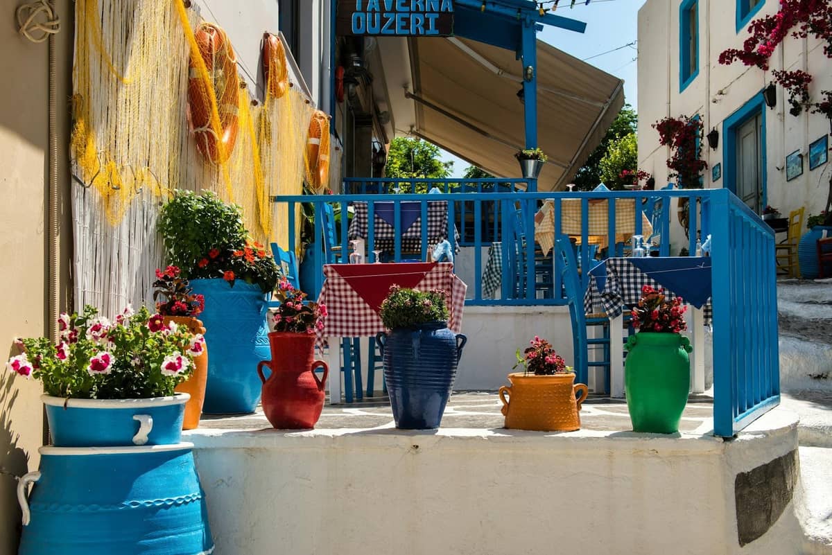 Encantadora entrada de taberna con barandillas azules, coloridas macetas y mesas dispuestas en una pintoresca calle encalada en Creta.