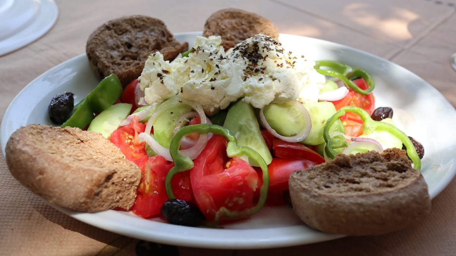 Um prato de dakos cretense tradicional coberto com tomates, pepinos, pimentões verdes, azeitonas e queijo cremoso, rodeado de torradas de cevada, sobre uma mesa ao ar livre com luz solar.