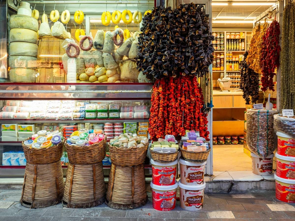 Una tienda con verduras secas colgantes, quesos, aceite de oliva, especias y productos de despensa en exhibición, evocando una escena de mercado en Creta.