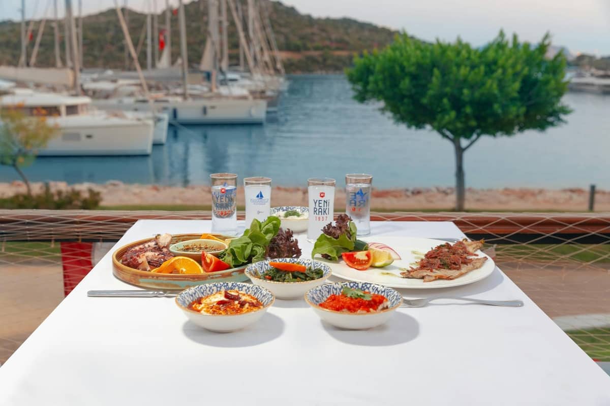 Mesa servida con platos cretenses, copas de raki y vistas a una marina con barcos y colinas al fondo.
