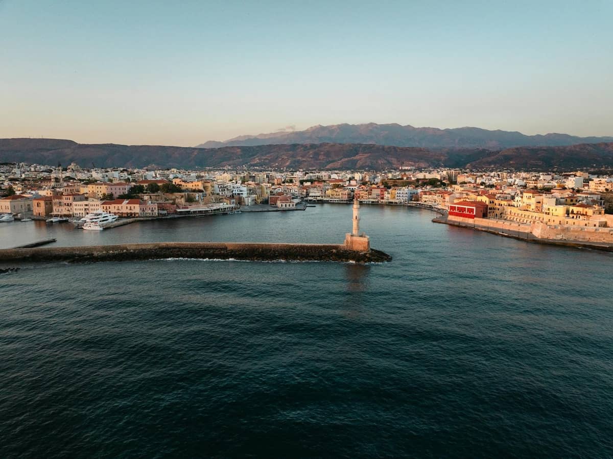 Vista aerea del porto veneziano di Chania, Creta, con un faro, edifici sul lungomare e montagne sullo sfondo.