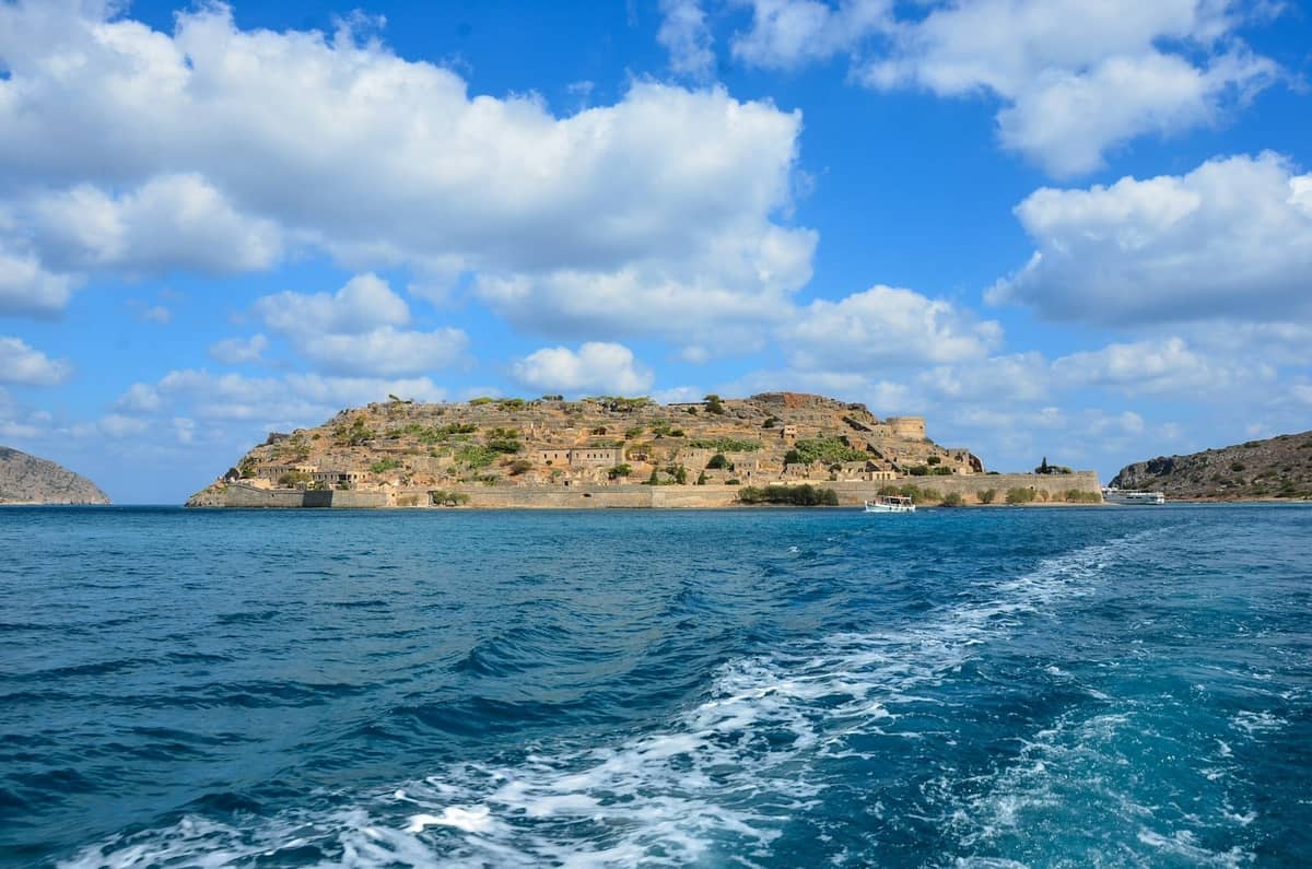 Vista dell'isola di Spinalonga dal mare in una giornata di sole, con la scia di una barca che conduce verso la storica fortezza dell'isola.