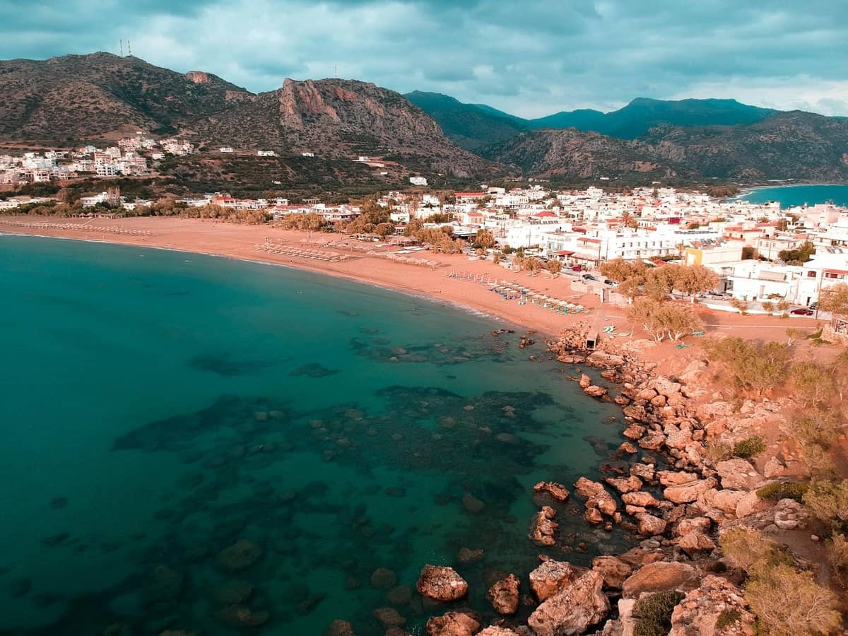 Luftaufnahme einer Küstenstadt auf Kreta mit klarem türkisfarbenen Wasser, felsiger Küstenlinie, Strand und Bergen im Hintergrund.