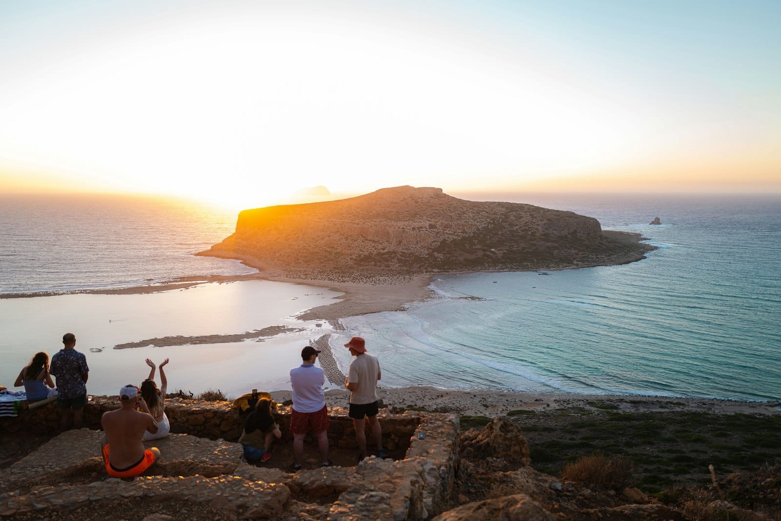 Casal assistindo a um pôr do sol deslumbrante sobre a lagoa de Balos em Creta, com o mar, os penhascos e a ilha iluminados por uma luz dourada, criando uma atmosfera romântica.