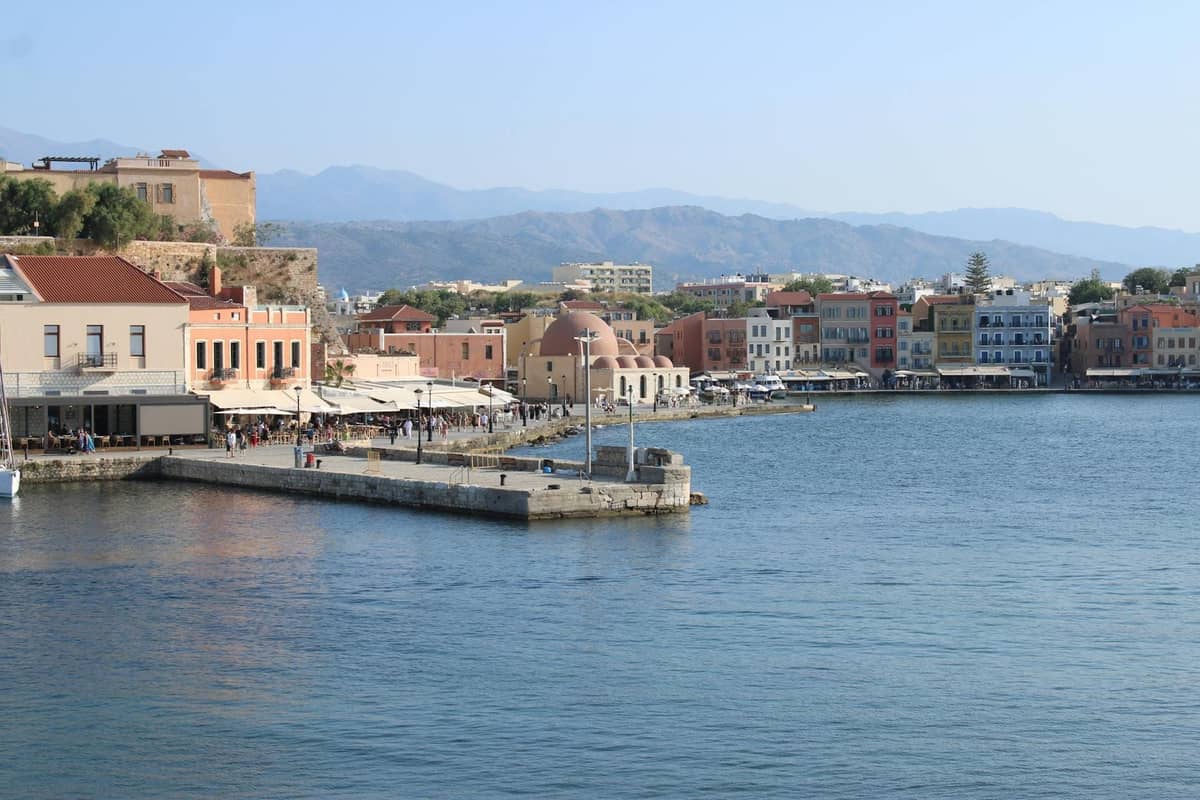 Vista costera del casco antiguo de Chania con edificios coloridos, paseo marítimo y montañas lejanas bajo un cielo despejado.