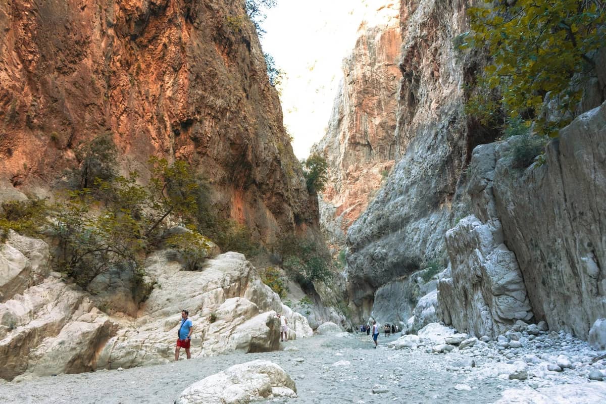 Amplia garganta en Creta con excursionistas caminando por el lecho rocoso del río, rodeados de altas paredes de cañón y escasos árboles otoñales.