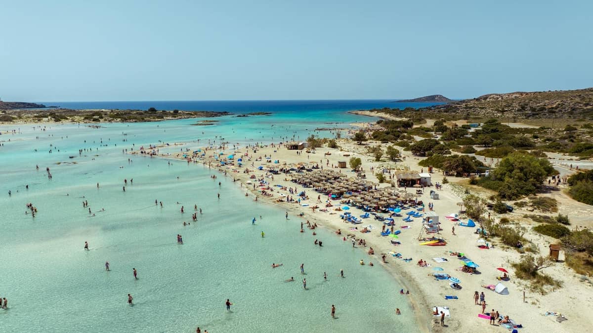 Vista aerea di una spiaggia popolare a Creta con acque turchesi poco profonde, bagnanti, ombrelloni e persone che nuotano e si rilassano lungo la riva sabbiosa.