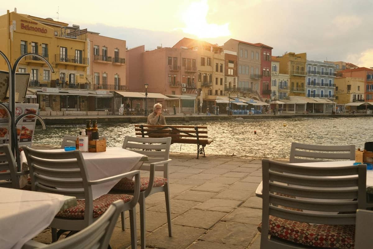 Tavoli vuoti di ristorante all'aperto al tramonto nel Porto Veneziano di Creta, con edifici colorati e acqua sullo sfondo.