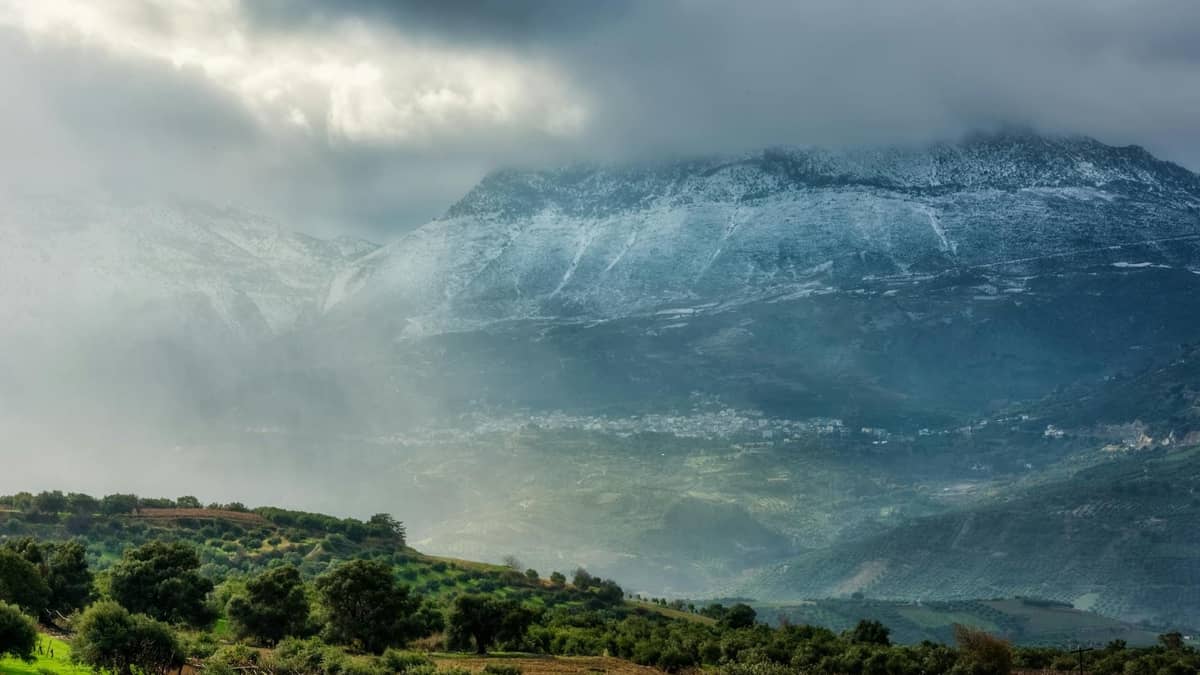 Vista drammatica e nuvolosa delle Montagne Bianche con vette innevate, colline lussureggianti e case di villaggio sparse nell'entroterra occidentale di Creta