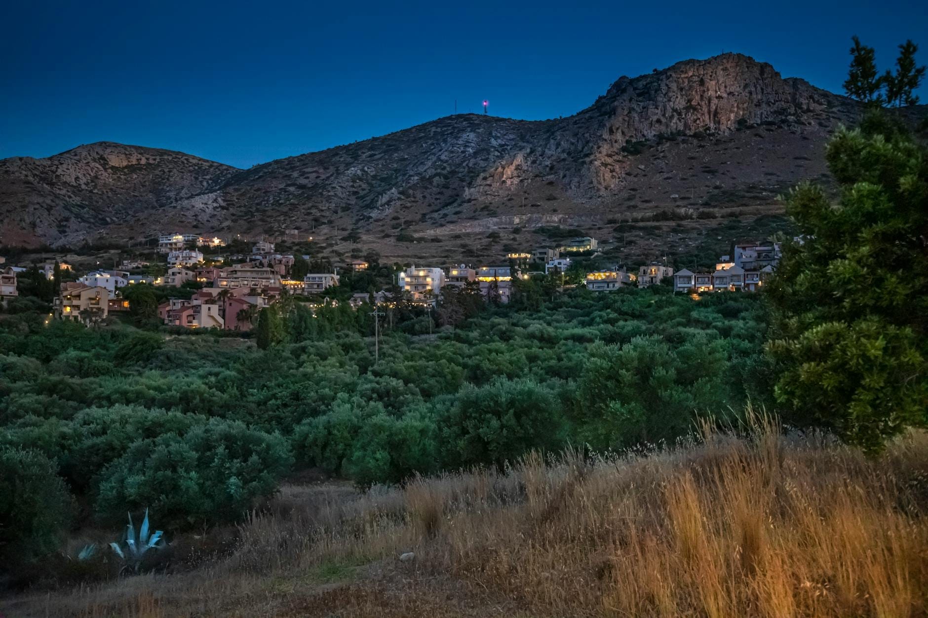 Vila de montanha em Creta ao entardecer, com casas aninhadas entre oliveiras e colinas rochosas ao fundo, sob um céu de crepúsculo profundo.