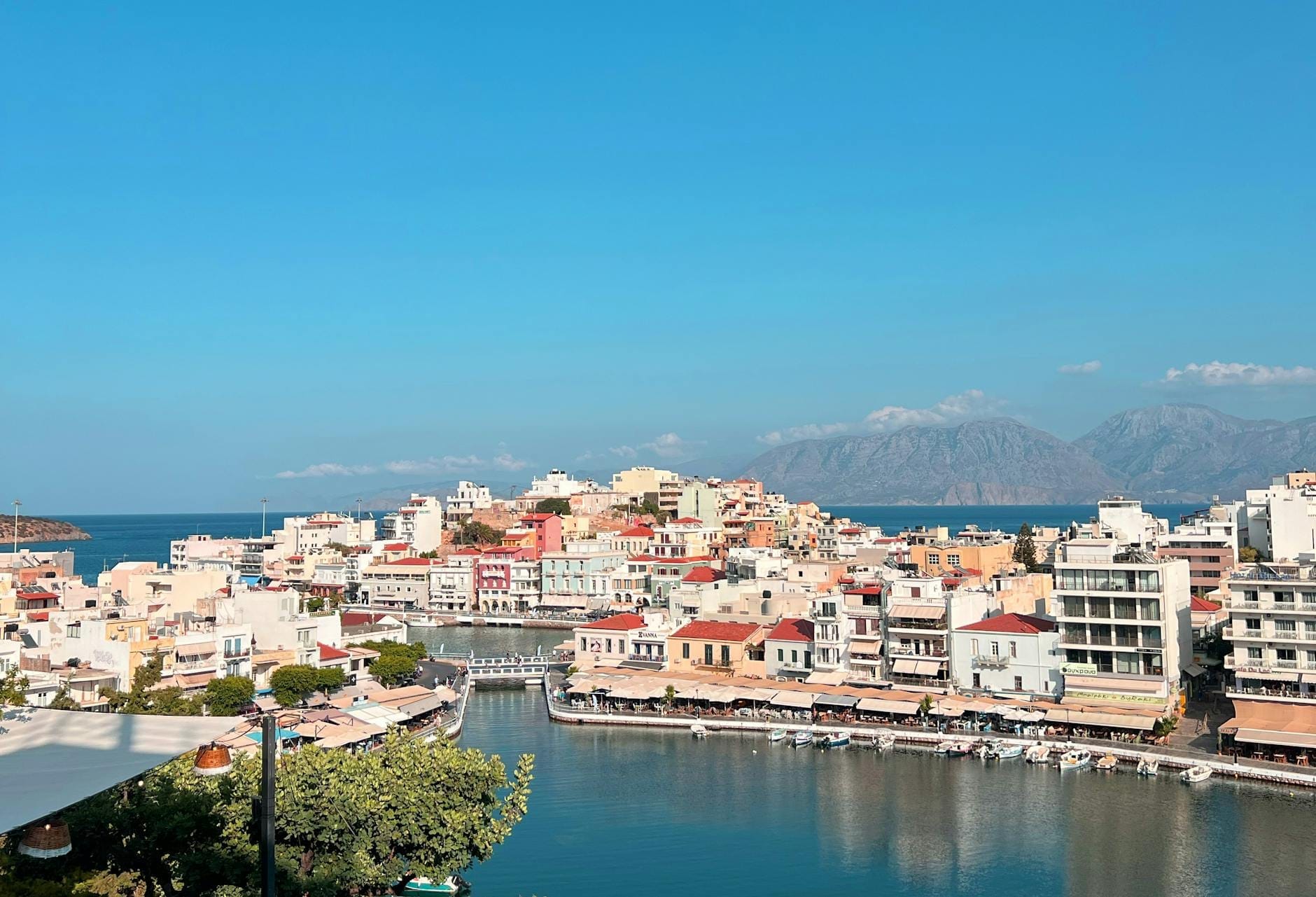 Vista panorâmica de uma colorida cidade cretense com construções tradicionais à beira-mar, montanhas ao fundo e céu azul limpo.