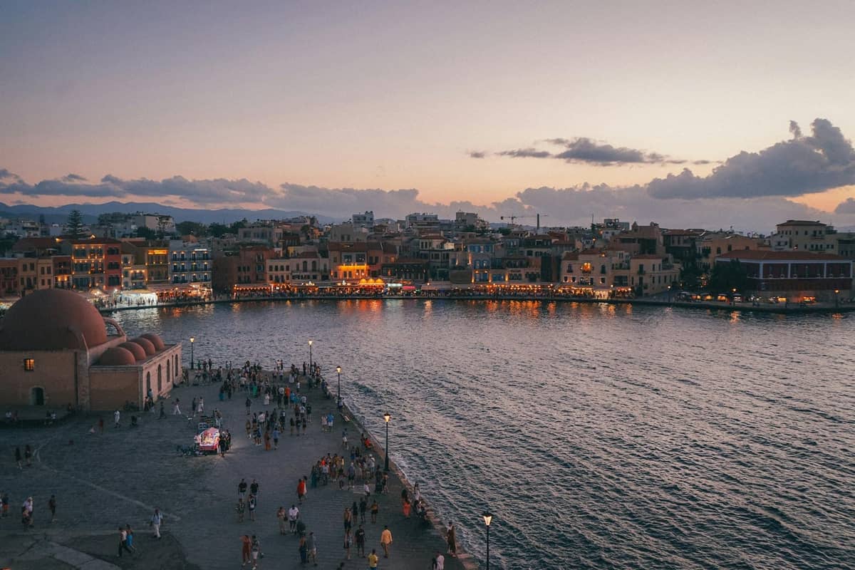 Vista noturna do Porto Veneziano de Chania em Creta, com edifícios da cidade antiga, calçadão à beira-mar e multidões aproveitando a atmosfera animada ao pôr do sol.