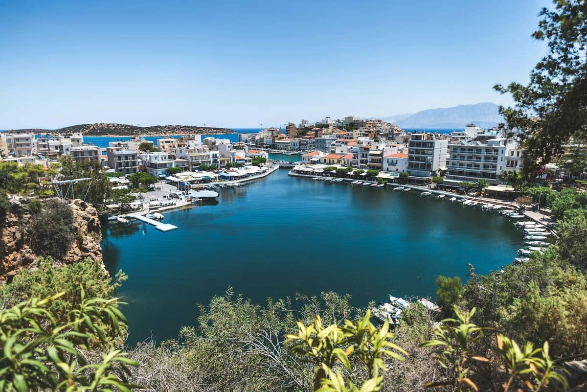 Um lago portuário ensolarado rodeado de edifícios e barcos em Agios Nikolaos, Creta, com o mar e as montanhas ao fundo.