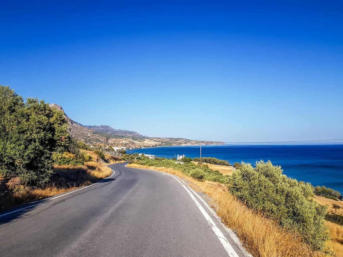 Estrada costeira sinuosa em Crete com o mar azul à direita, grama dourada e colinas sob um céu azul limpo.