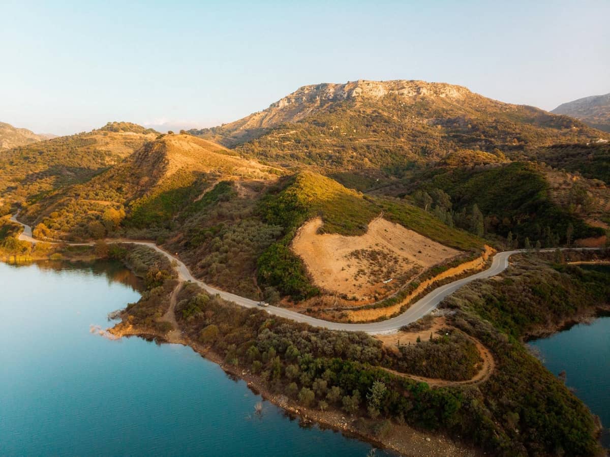Estrada costeira sinuosa em Crete com água turquesa, colinas verdejantes e um pano de fundo montanhoso sob céus limpos.