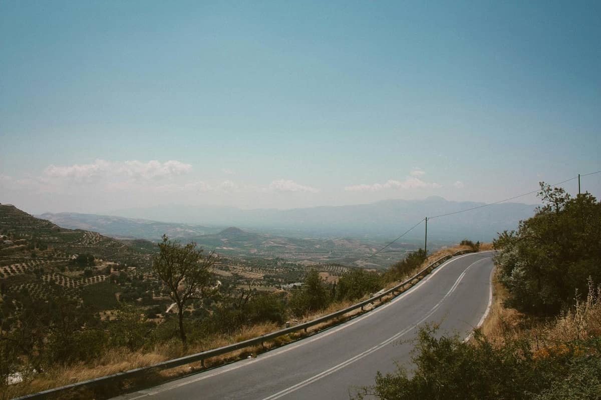 Estrada sinuosa com grades de proteção por uma paisagem montanhosa aberta, com vista para vales e montanhas distantes sob um céu azul limpo em Creta.