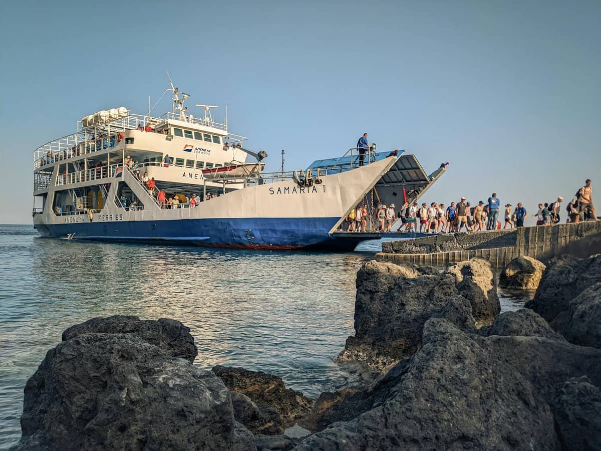 Pasażerowie wsiadający na duży prom przy nabrzeżu pod bezchmurnym niebem w śródziemnomorskiej scenerii, z skalistym brzegiem na pierwszym planie.