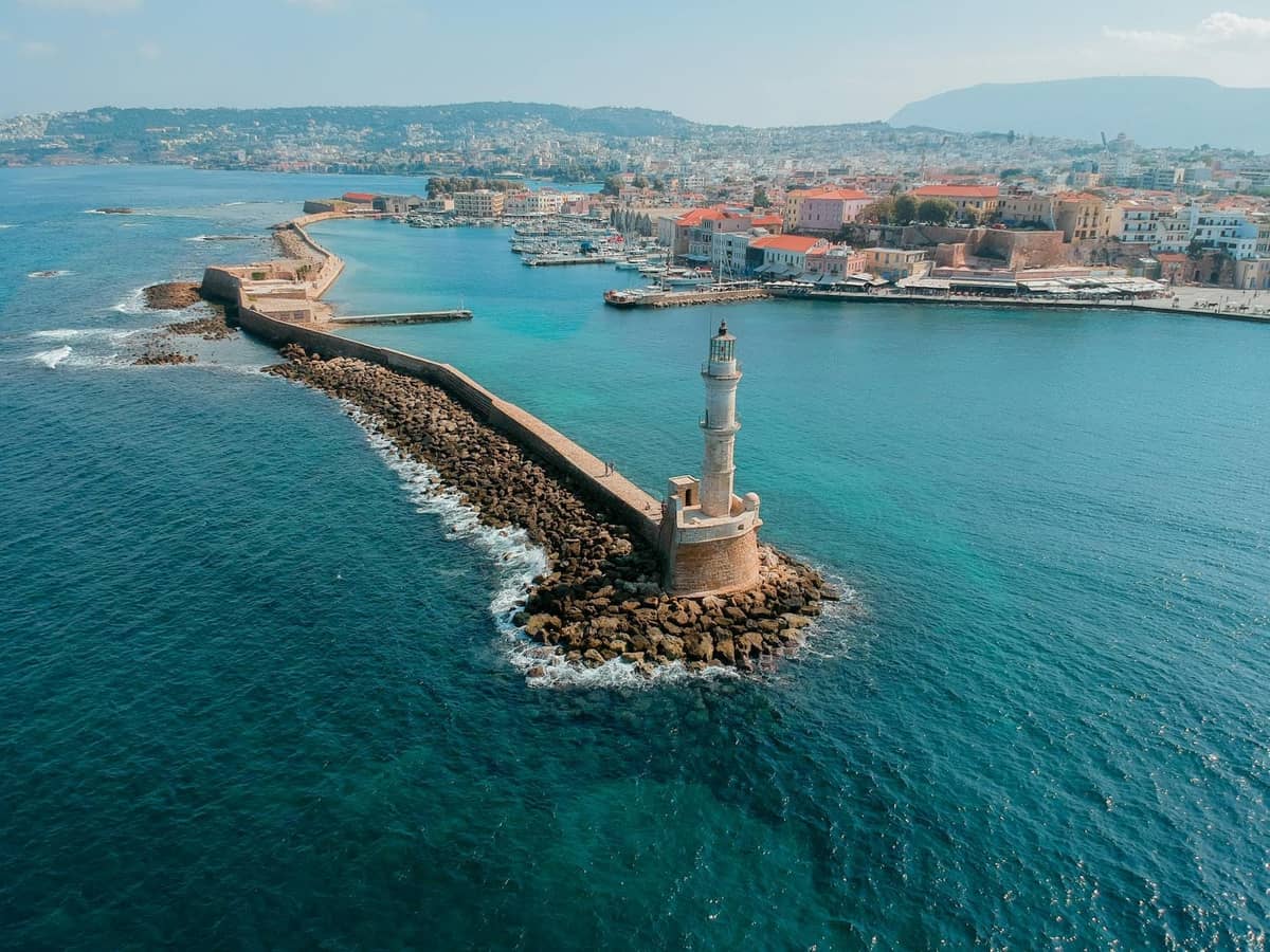 Vista aerea del porto di Chania a Creta, con il caratteristico faro, il lungo molo e il colorato centro storico con le montagne sullo sfondo.