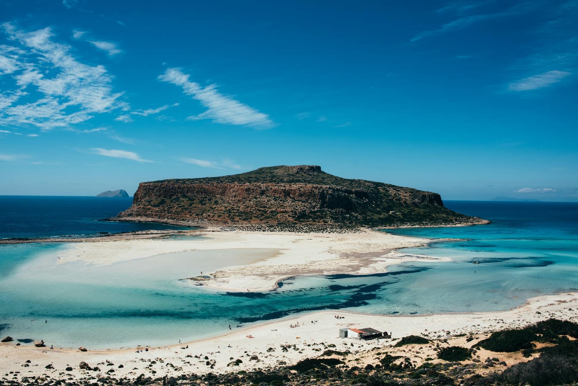 Vista panorâmica deslumbrante de uma lagoa ensolarada com águas turquesa e uma grande ilha rochosa, emoldurada por céu azul e praias de areia na Grécia.
