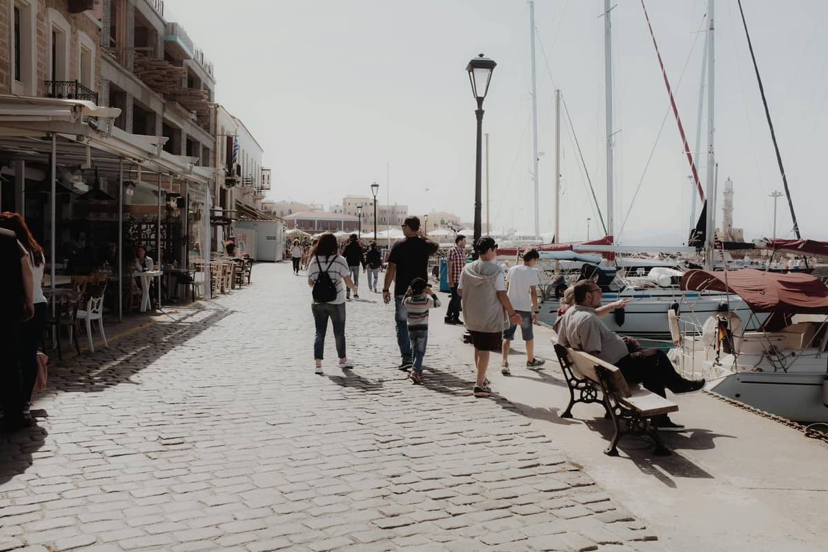 Keluarga dan pengunjung berjalan-jalan di sepanjang promenade pelabuhan yang indah di Krete, dengan perahu yang berlabuh dan kafe-kafe yang berjajar di sepanjang jalan berbatu.