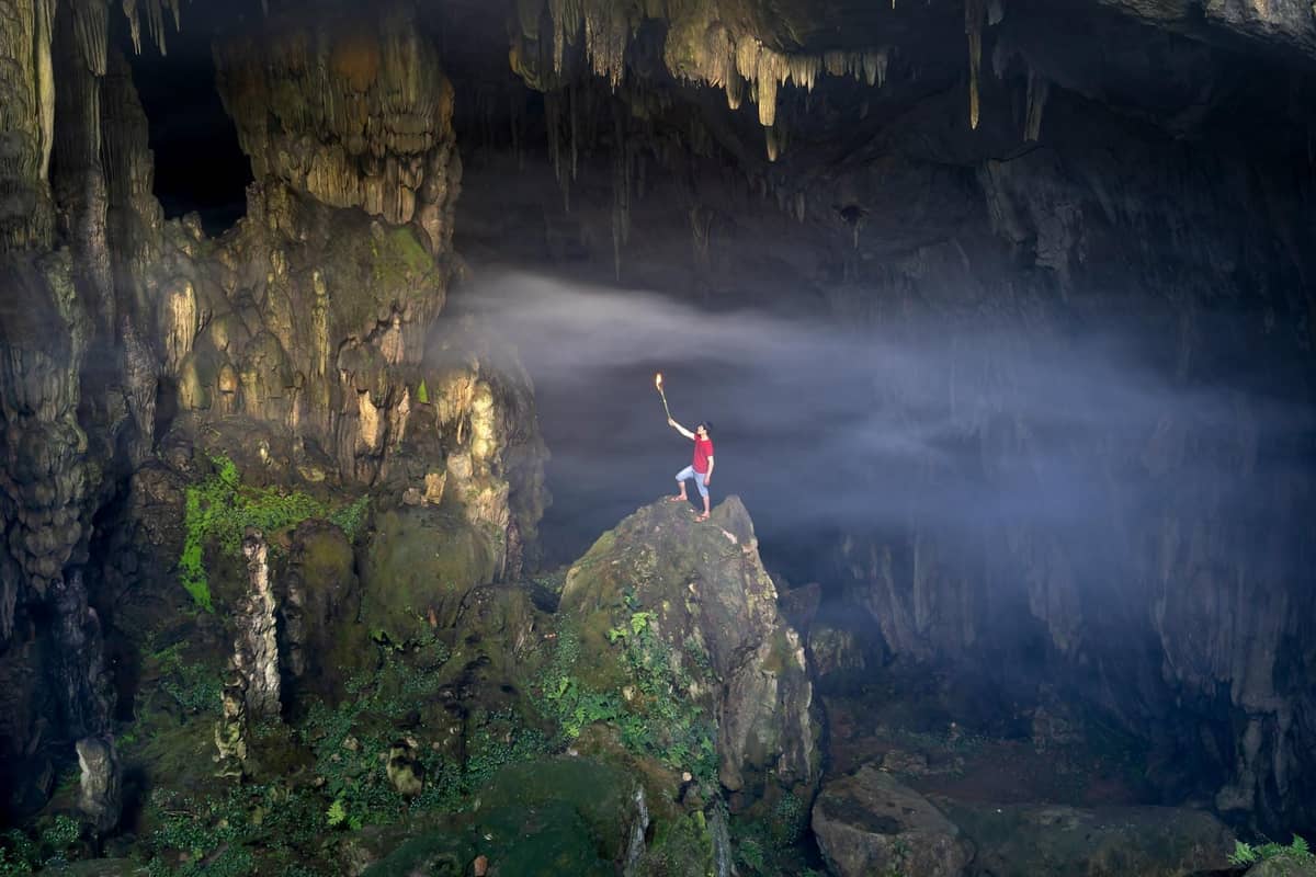Seseorang memegang obor di dalam gua besar dengan stalaktit, stalagmit yang dramatis dan seberkas sinar matahari yang menerangi bagian dalam gua.