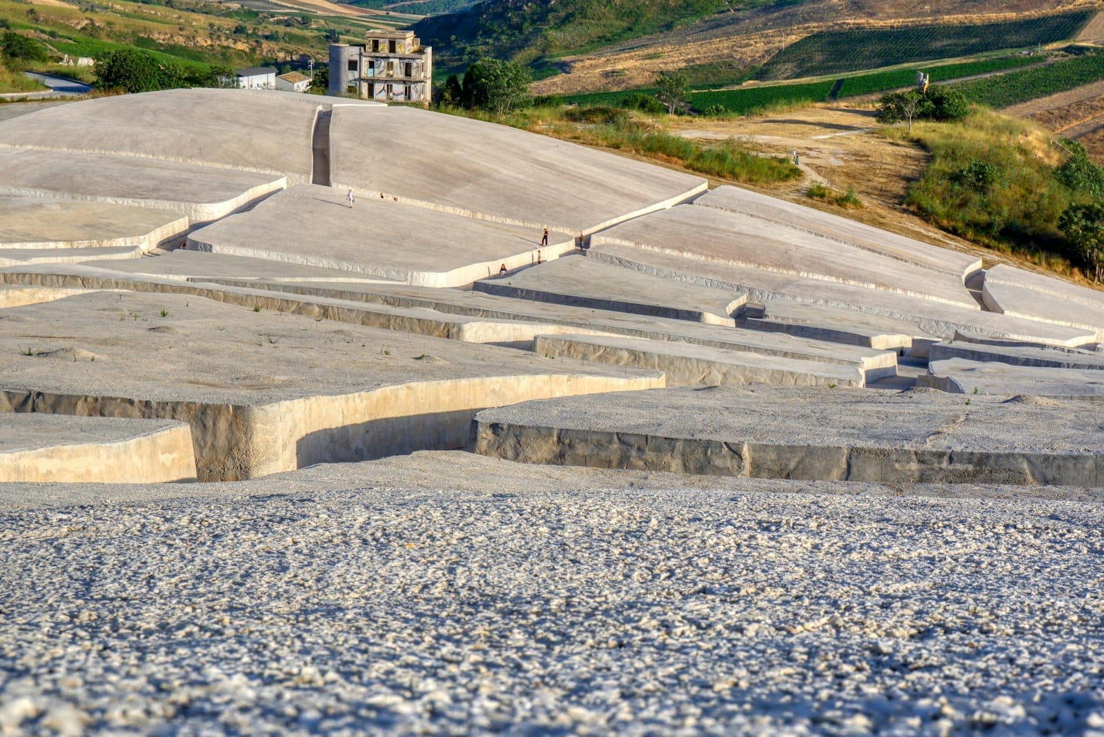 Weitläufiger Blick auf die weißen Betonblöcke des Cretto di Burri mit vereinzelten Besuchern und einem verlassenen Gebäude in der sizilianischen Landschaft.