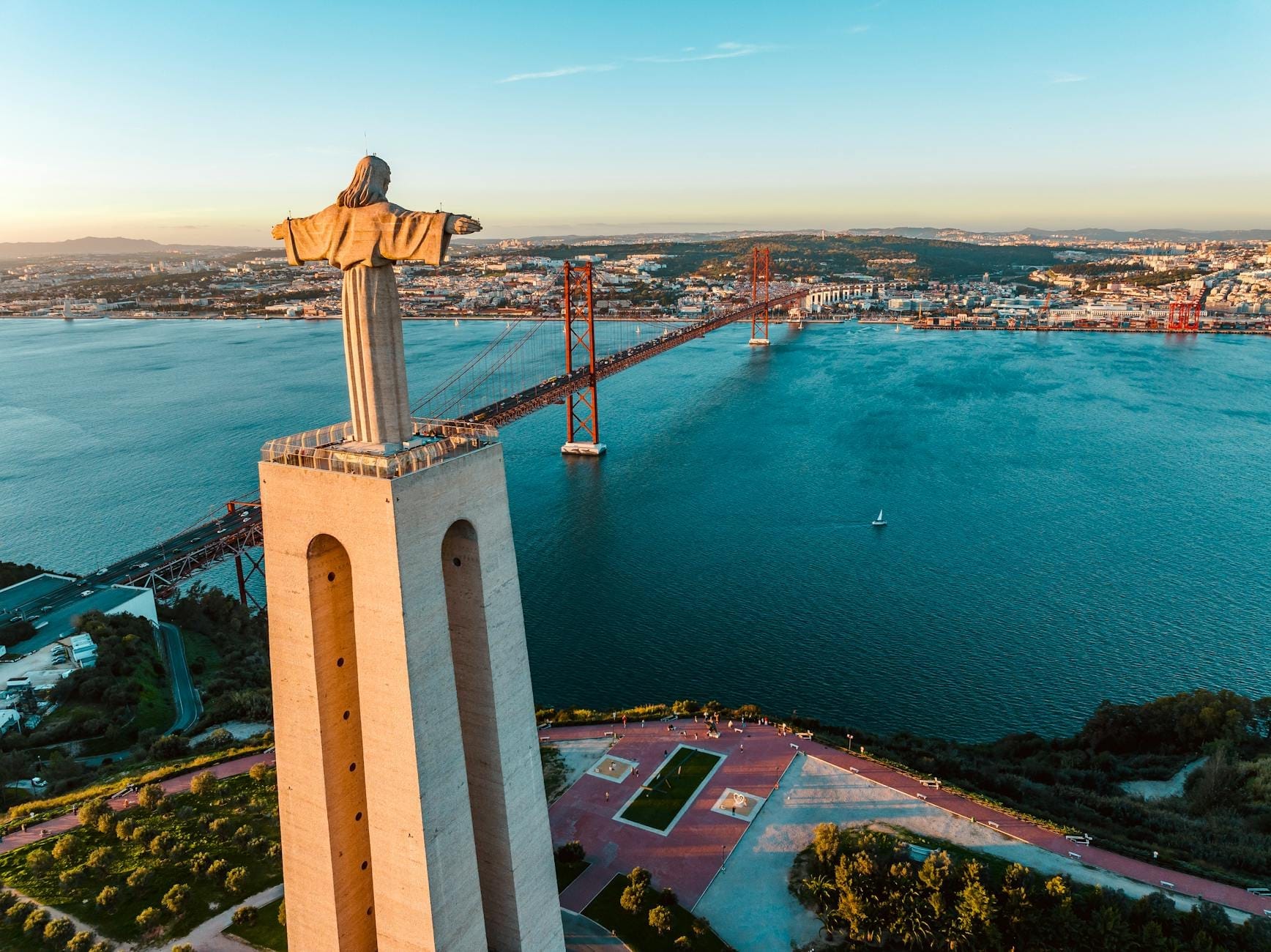 Vista aerea della statua di Cristo Rei che domina il Tago e il Ponte 25 de Abril, con Lisbona e il paesaggio circostante sotto un cielo azzurro e limpido.