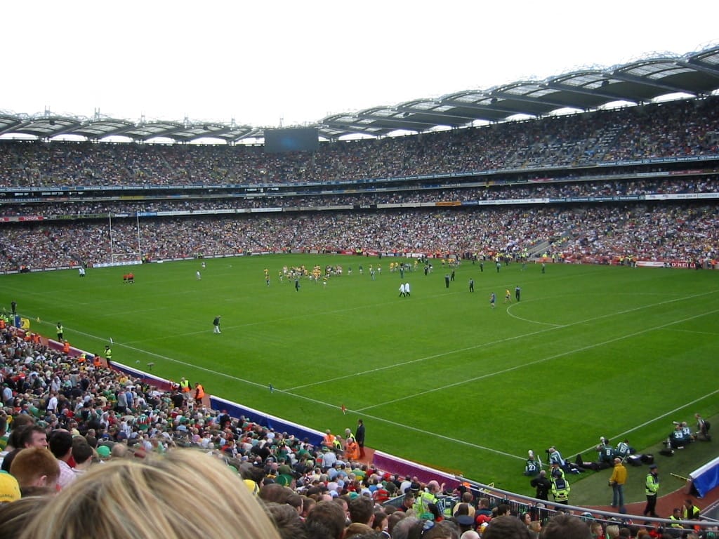 Lo stadio Croke Park gremito di migliaia di spettatori durante una partita di giochi gaelici, campo verde lussureggiante sotto un cielo nuvoloso, atmosfera vibrante dello stadio.