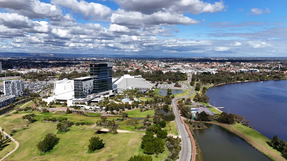 Aerial view of Crown Perth entertainment complex with its distinctive hotel towers, lush green grounds, and the Swan River running alongside on a partly cloudy day.
