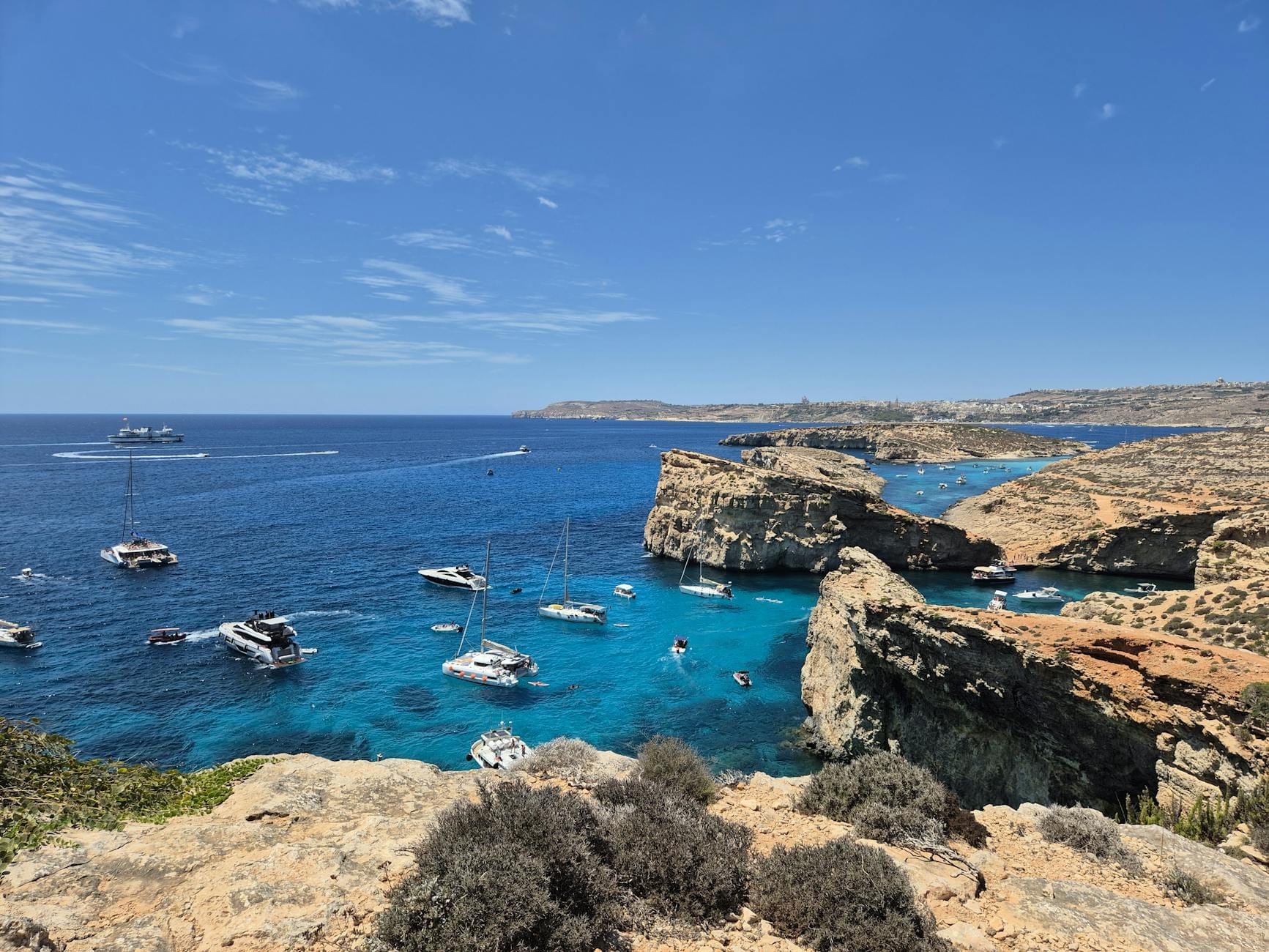Vista spettacolare della Crystal Lagoon con acque turchesi, scogliere rocciose e diverse barche ormeggiate nella baia tranquilla sotto un cielo azzurro limpido.