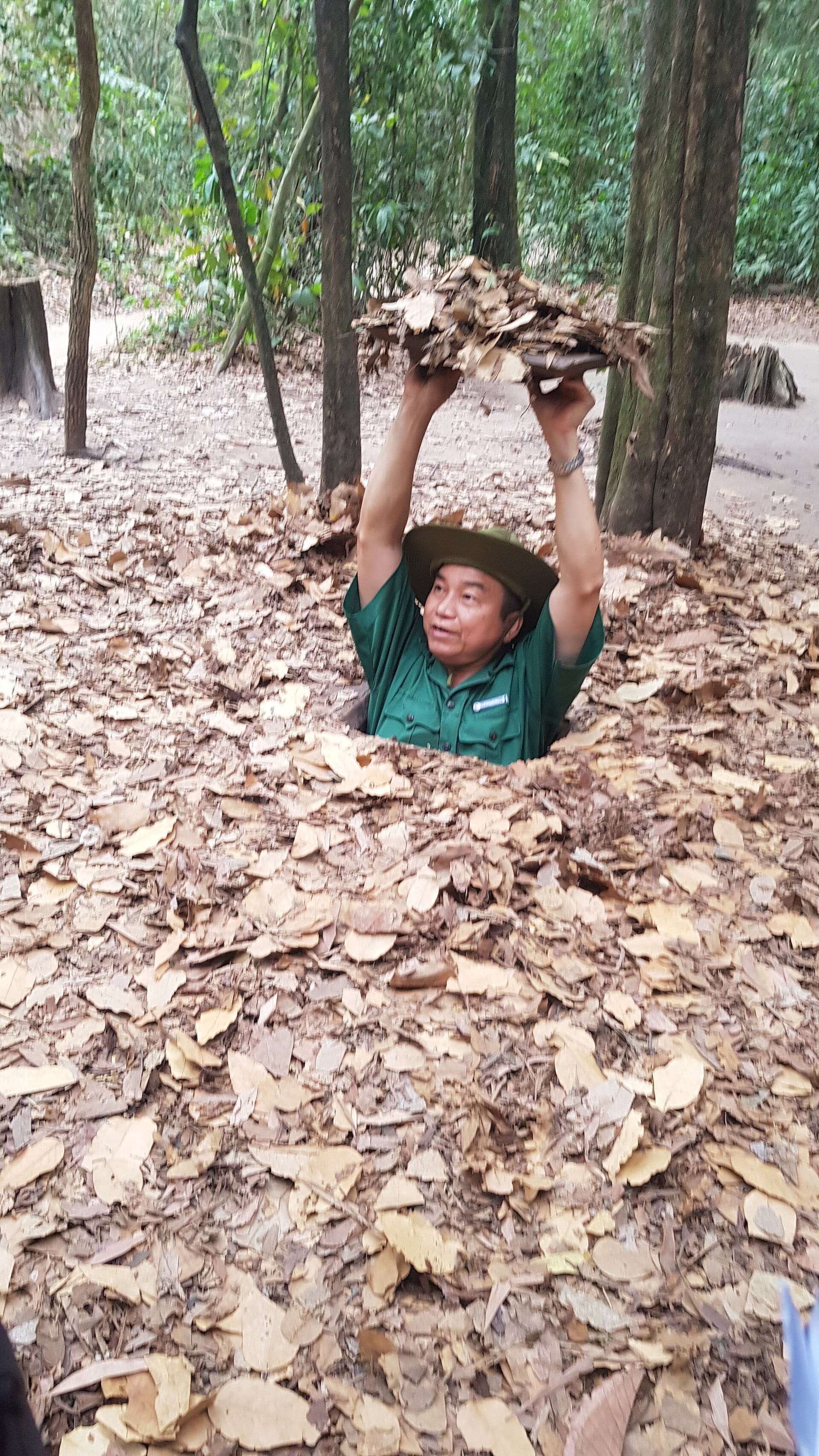 Guía emergiendo de una entrada de túnel oculta cubierta de hojas en los Túneles de Củ Chi, rodeada de densa vegetación y suelo terroso.