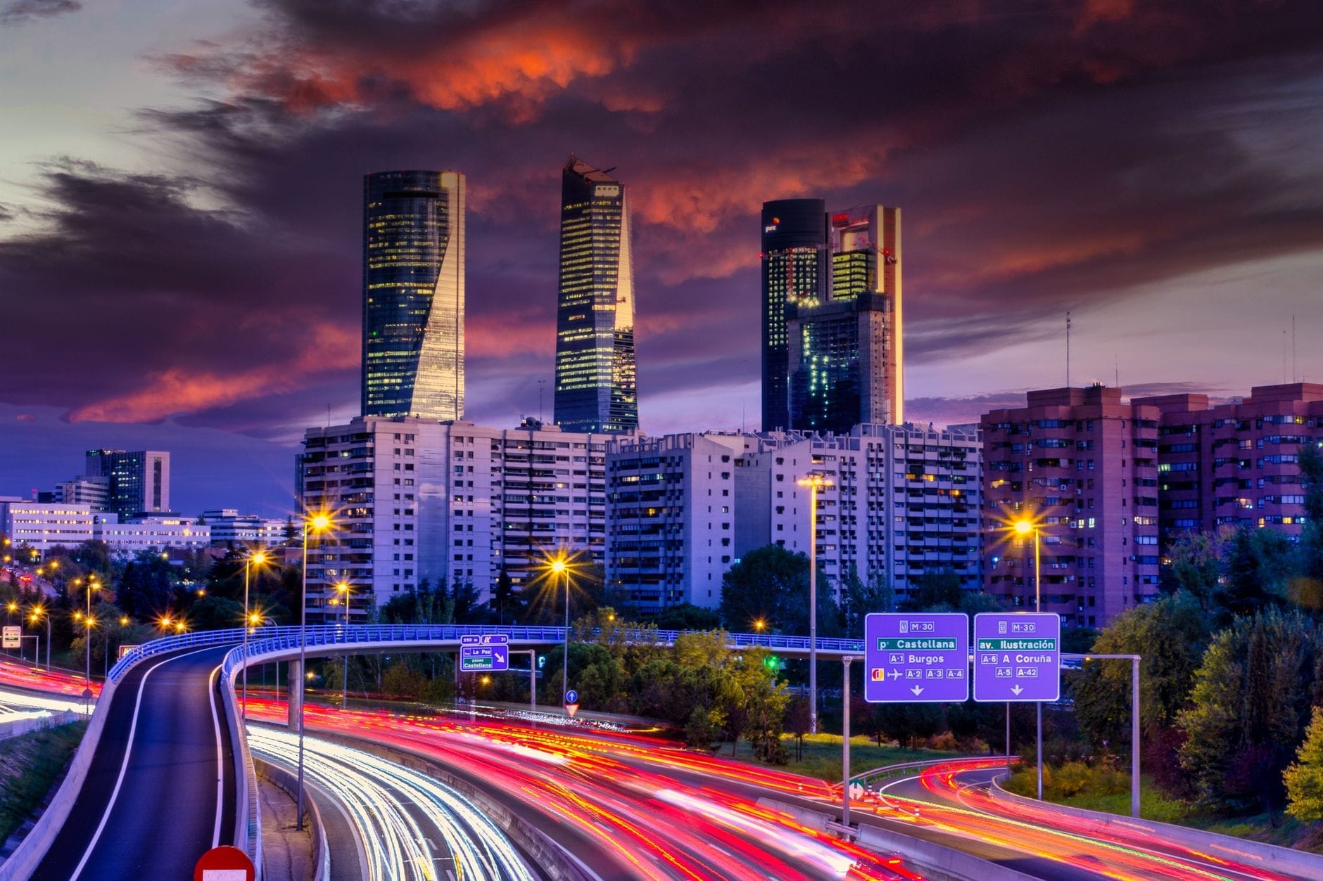 I grattacieli Cuatro Torres al tramonto, con nuvole spettacolari in cielo e le strade illuminate con scie di luce in primo piano, skyline di Madrid.