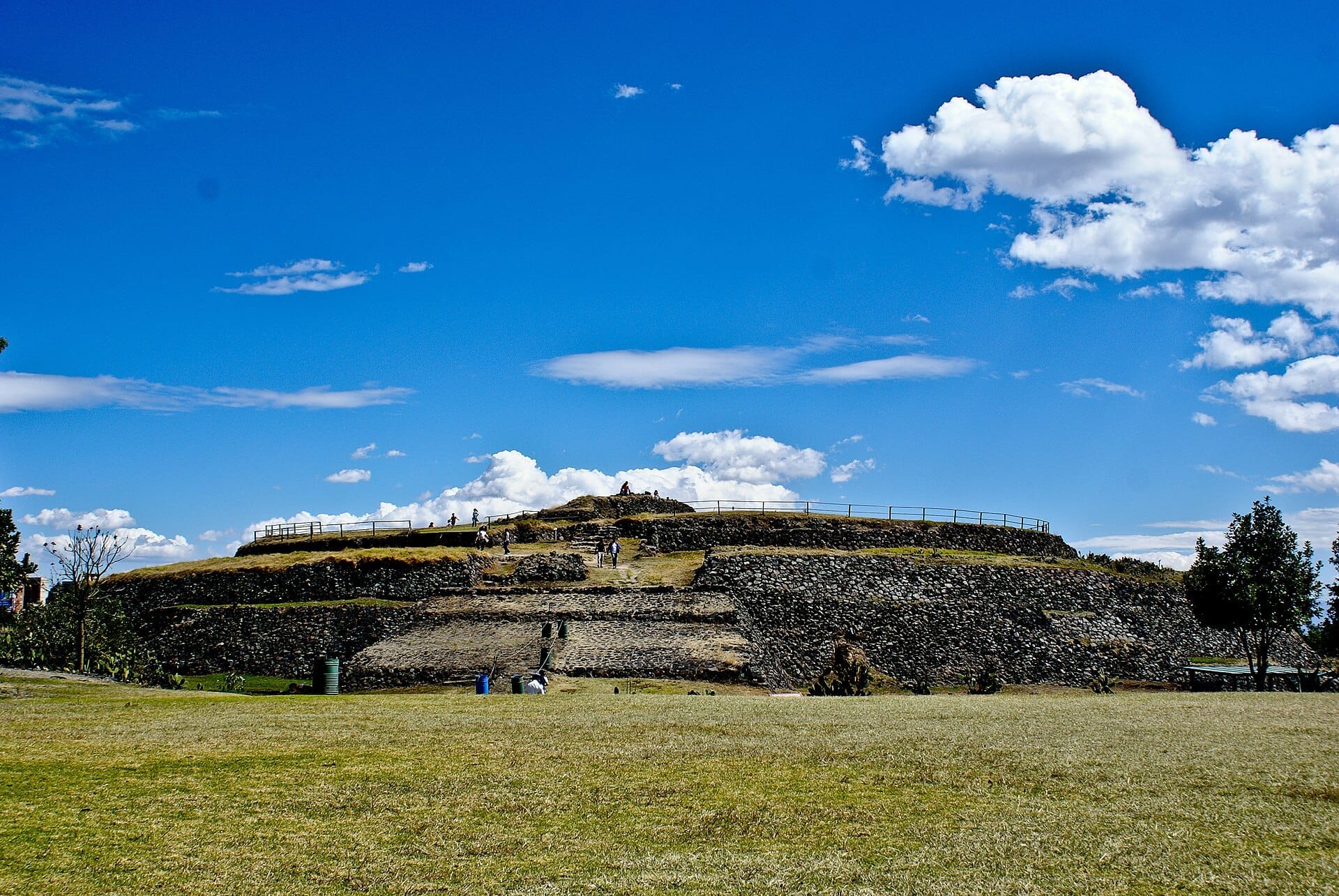 Inquadratura ampia della piramide circolare nella Zona Archeologica di Cuicuilco, con terrazze in pietra, prati e visitatori sotto un cielo azzurro brillante.
