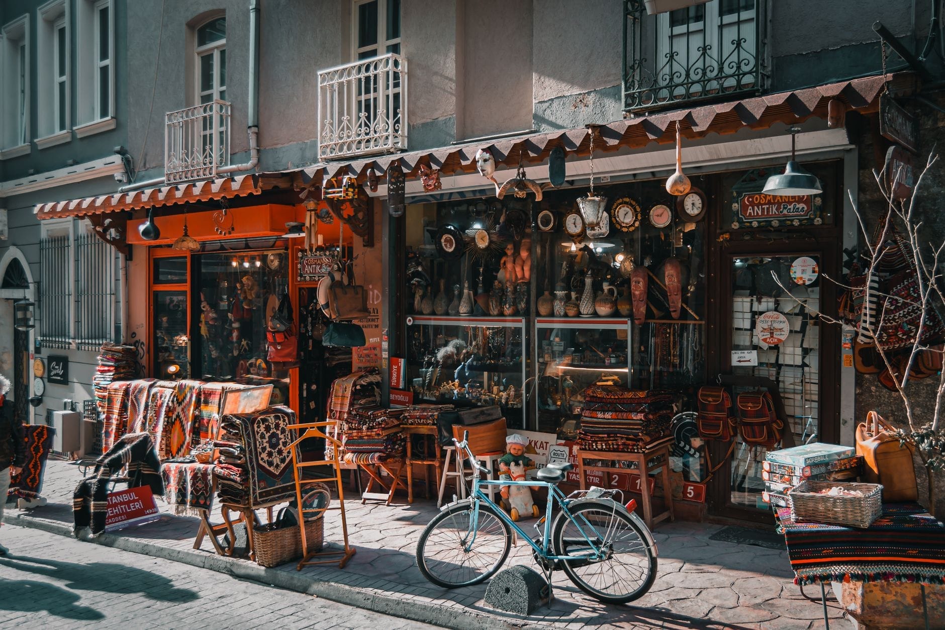 Colorato negozio di antiquariato nel Quartiere delle Antichità di Çukurcuma, Istanbul, con tappeti impilati, oggetti vintage e una bicicletta color verde acqua su una strada acciottolata.