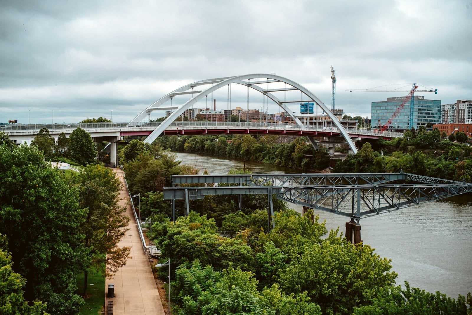 Vista del sendero peatonal en Cumberland Park en Nashville, con exuberante vegetación, el río y un moderno puente arqueado bajo cielos nublados.