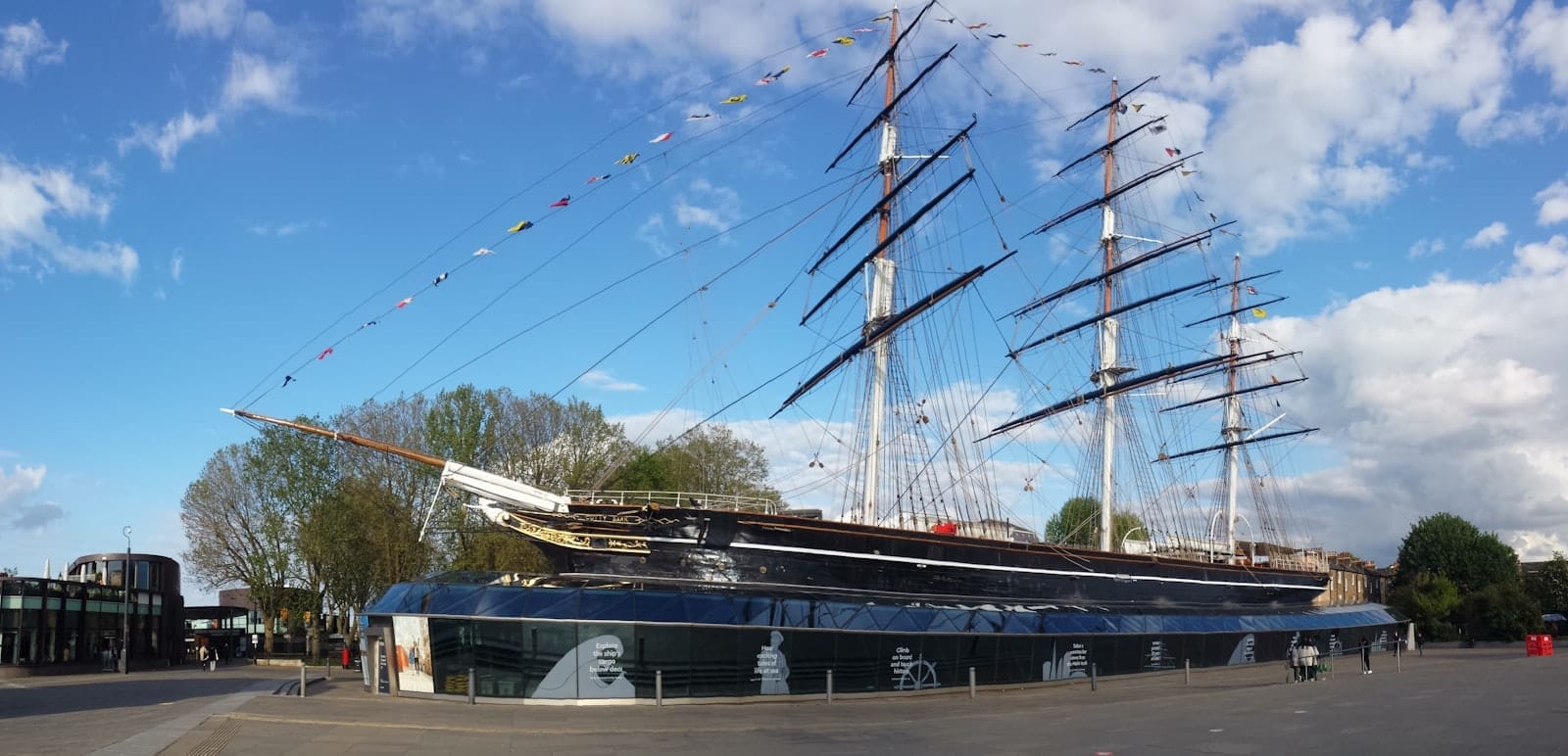 Lo storico clipper da tè Cutty Sark ormeggiato a Greenwich, circondato da una piazza di vetro, sotto un cielo azzurro brillante con nuvole sparse.