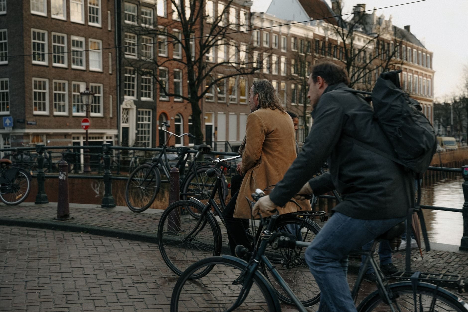 Dos ciclistas cruzando un puente sobre un canal en Ámsterdam, rodeados de bicicletas aparcadas y edificios históricos junto al canal.
