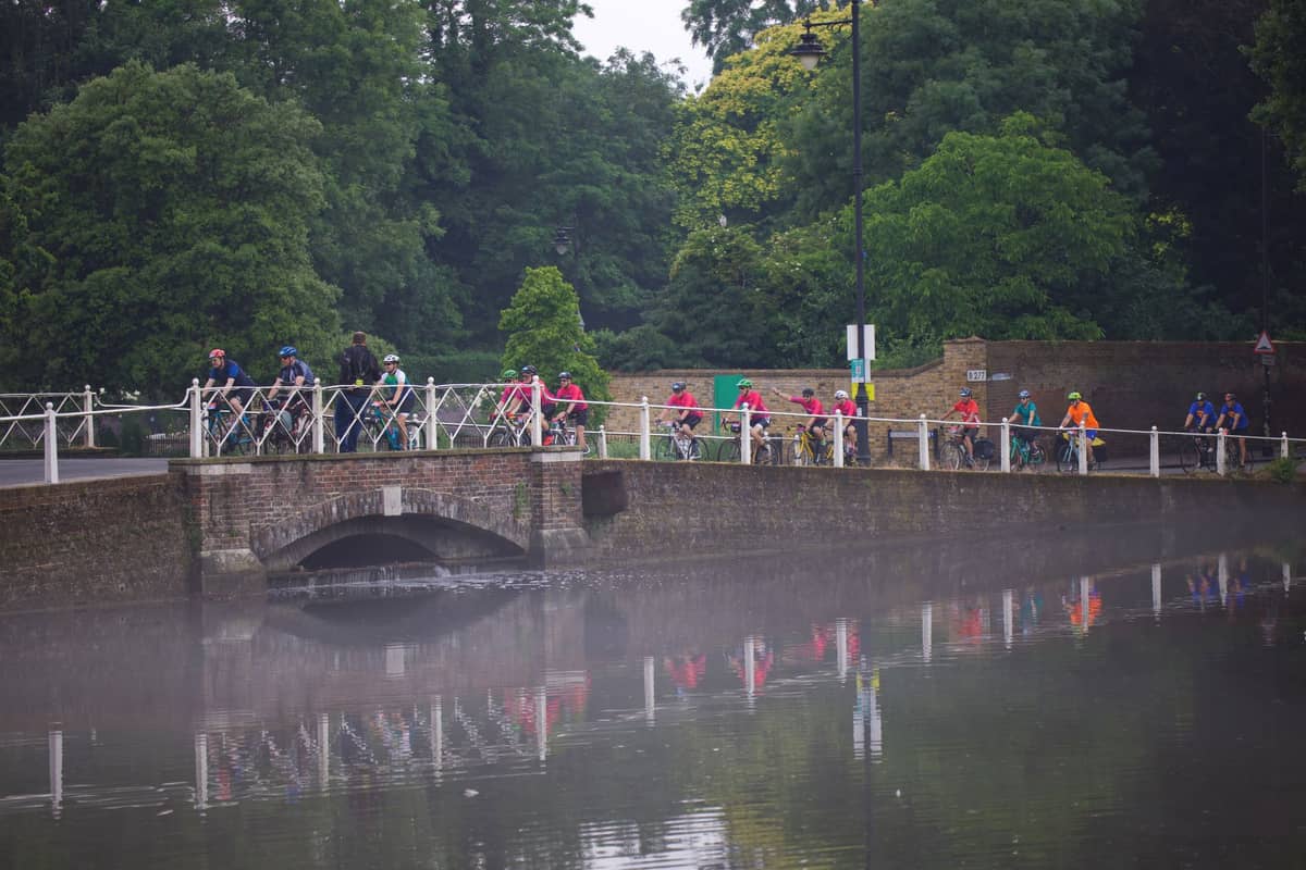 Cyclists ride across a brick bridge over a canal in a green, park-like setting with mist above the water, evoking Bruges' flat, bike-friendly landscape.