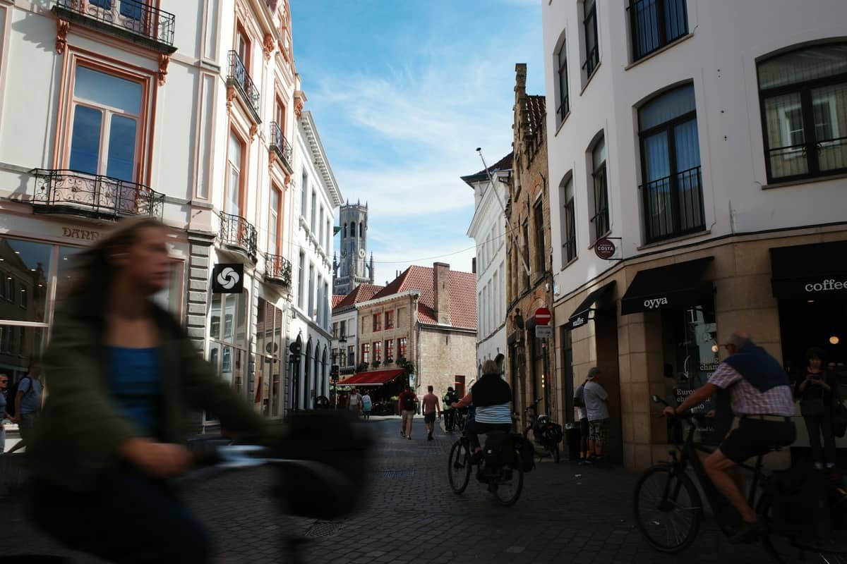 Cyclists ride through a cobblestone street in Bruges’ historic center, with the Belfry of Bruges visible in the background under a blue sky.
