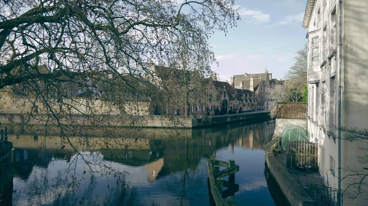 View of a picturesque canal flanked by historic Bruges buildings and tree branches, under a partly cloudy sky.