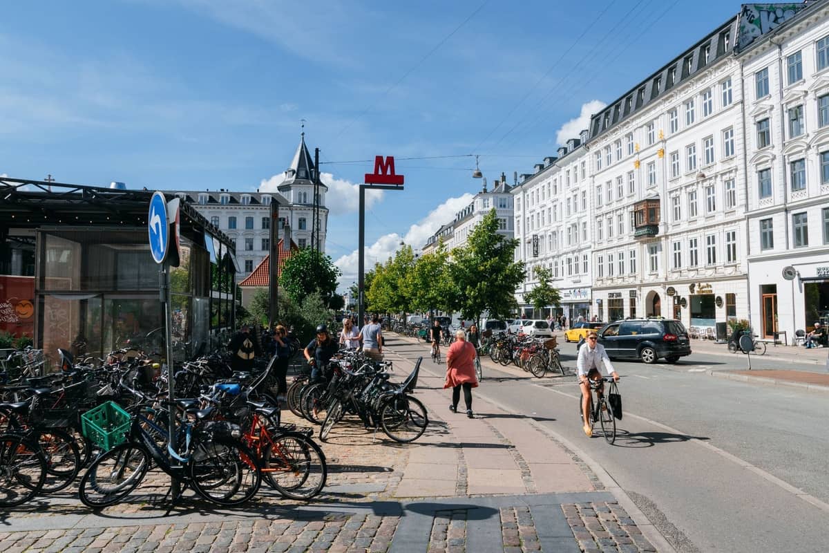 Ruchliwa ulica miejska w Kopenhadze z wieloma zaparkowanymi rowerami, rowerzystami na ścieżce rowerowej i klasyczną duńską architekturą w tle.