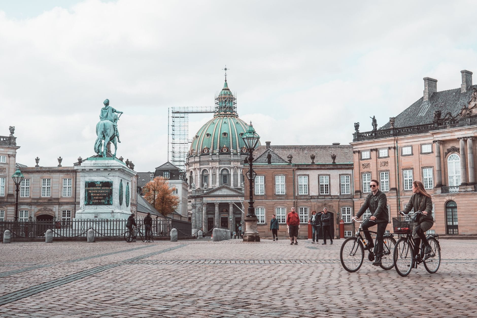 Duas pessoas pedalando por uma praça aberta em Copenhague com a Igreja de Mármore e prédios históricos ao fundo em um dia nublado.