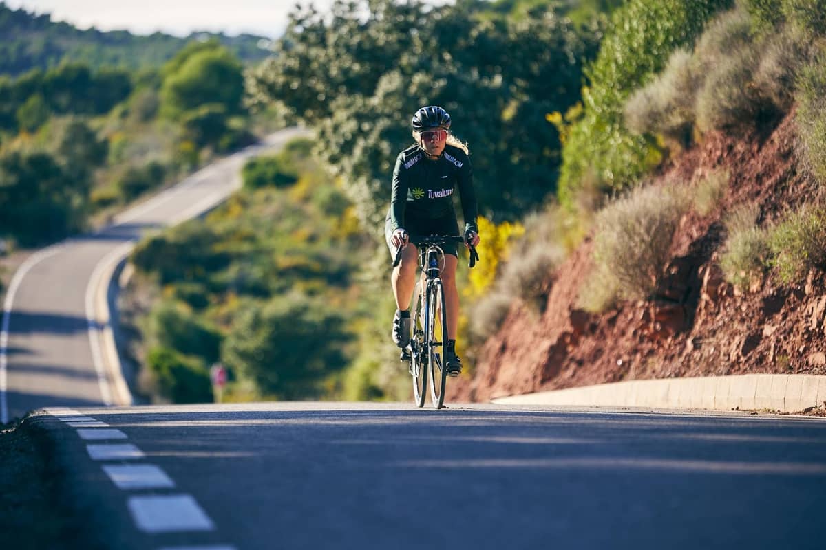 Cycliste roulant sur une route sinueuse bordée de végétation méditerranéenne et de douces collines, sous un ciel clair et ensoleillé.