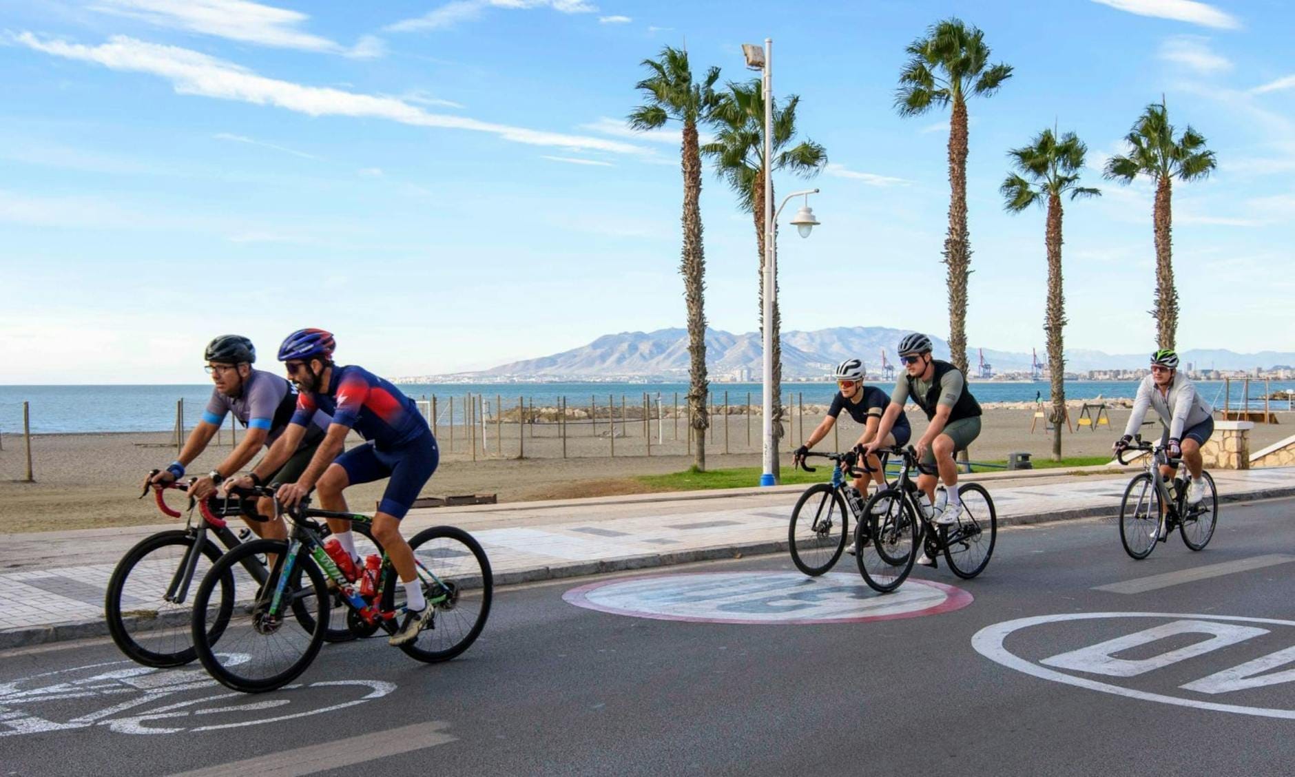 Grupo de ciclistas pedalando ao longo de uma estrada costeira ladeada de palmeiras em Mallorca com montanhas e mar azul ao fundo sob céu limpo.
