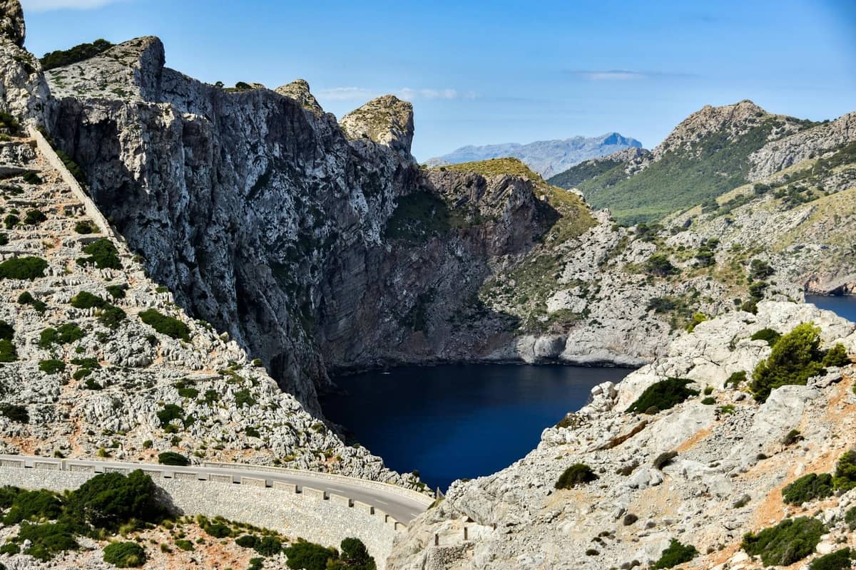 Une route de montagne sinueuse et pittoresque longe un littoral rocheux spectaculaire avec des falaises abruptes et une mer bleue, sous un ciel lumineux à Majorque.
