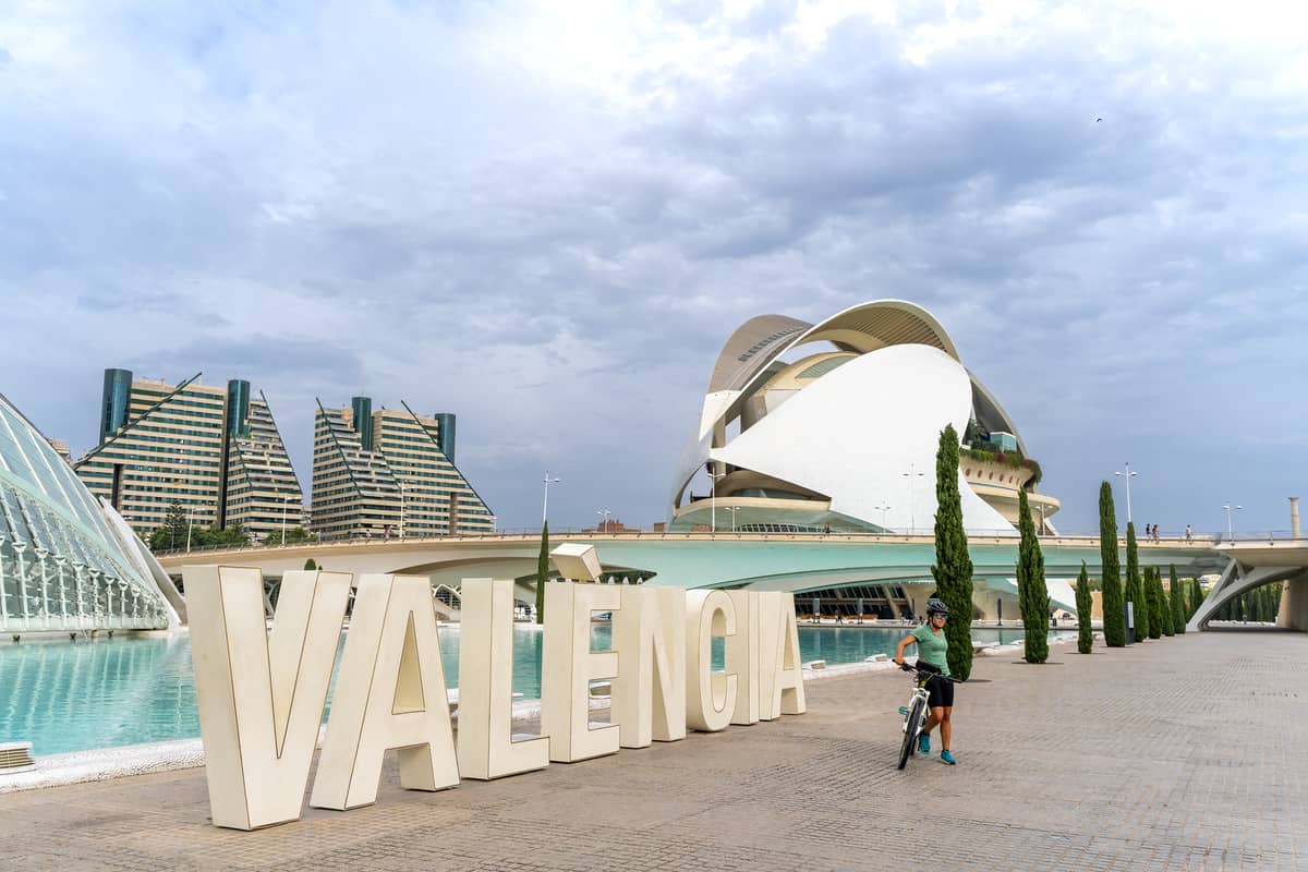 Ciclista in posa davanti all'insegna VALENCIA alla Città delle Arti e delle Scienze, con l'architettura modernista e le vasche riflettenti sullo sfondo in una giornata nuvolosa.
